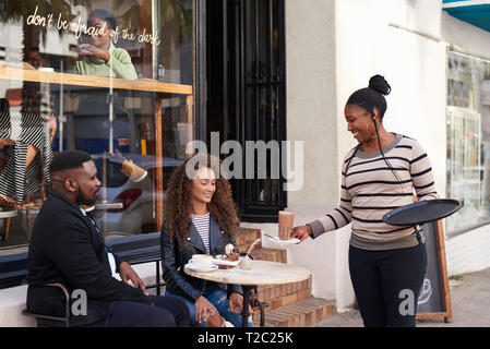 Gentile cameriera che serve drinks per sorridere giovani amici seduti insieme ad un tavolo esterno a un cafè sul marciapiede Foto Stock