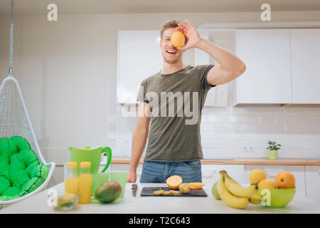 Sorridente giovane uomo bello detiene arancio dolce nella parte anteriore del viso mentre la cottura di frutta fresca in cucina. Cibo sano. Pasto vegetariano. La dieta detox Foto Stock