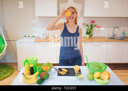 Sorridente giovane donna bionda detiene un pezzo di colore arancione nella parte anteriore del viso mentre la cottura di frutta fresca in cucina. Cibo sano. Pasto vegetariano. La dieta Foto Stock