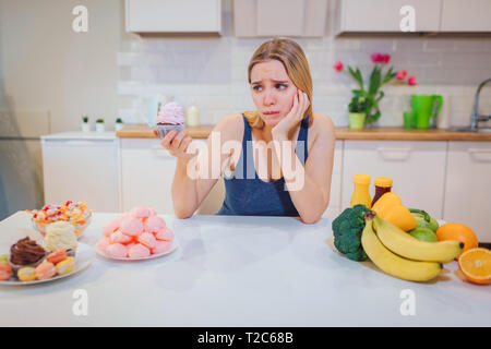 Giovane donna triste con torta dolce in una mano scegliendo tra un sano e malsano in cucina il cibo. Difficile scegliere tra frutta fresca Foto Stock