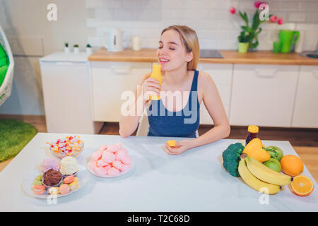 Giovane donna sorridente bevande acqua detox mentre scegliendo tra un sano e malsano in cucina il cibo. Difficile scegliere tra frutta fresca Foto Stock