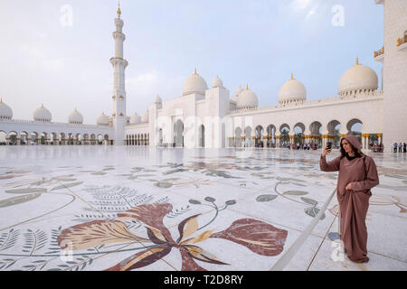 Giovane donna di scattare le foto alla Sheikh Zayed grande moschea cortile interno con ornati a tema floreale mosaici pavimentali, Abu Dhabi, Emirati Arabi Uniti Foto Stock