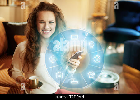 Felice giovane donna con lunghi capelli brunette con tazza da caffè utilizzando smart home application sullo smartphone e il robot vuoto piano di pulizia in background in Foto Stock