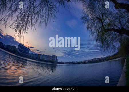 Mattina cityscape di scena a sunrise in primavera con un edificio residenziale che riflette nell'acqua e alcune nuvole in background e salici in Foto Stock