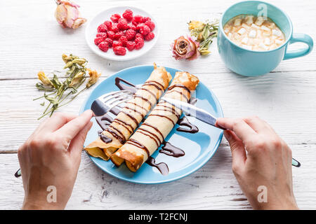 Foto di bianco tavolo in legno con frittelle ritorto versata con cioccolato, cup con il cacao, fiori secchi Foto Stock