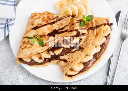 Crepes o blini farciti con cioccolato noccioletta, banana su una piastra bianca. Primo piano Foto Stock