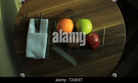 Un bicchiere coppa di frutta con Apple, Verde mela, arancio e Kiwi, con coltello e forchetta in igienico su una tavola di legno Tavolo rotondo Foto Stock