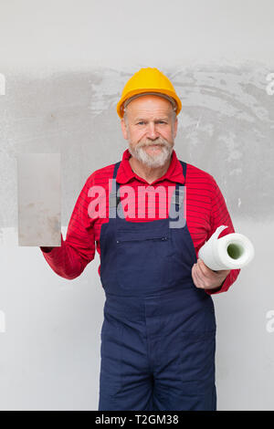 Costruttore di anziani con casco giallo azienda gli strumenti di pittura, che pongono di fronte a un muro di cemento - nuova casa in costruzione Foto Stock