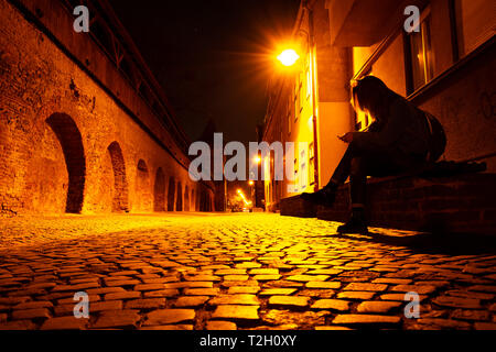 Giovane donna con il telefono in mano, su un banco a tarda notte, su uno stile medievale street a Sibiu in Romania, con ciottoli accesa caldo giallo/rosso da st Foto Stock