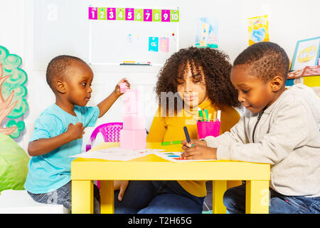 Due piccoli ragazzi neri con insegnante vivaio fare esercizi di classe di disegno e costruzione di blocchi Foto Stock