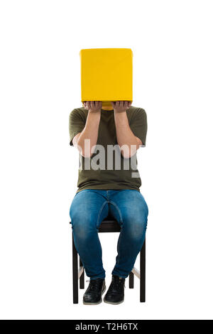 A piena lunghezza Ritratto di casual in incognito giovane uomo seduto su una sedia tenendo un riquadro giallo invece di testa isolata su bianco. Introverso persone nascondere Foto Stock