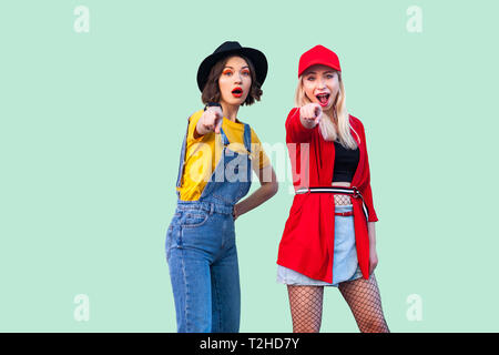 Migliori amici spendendo molto tempo insieme. Due belle stupiti di moda hipster ragazze in piedi e puntare il dito a voi, guardando la fotocamera. Indo Foto Stock