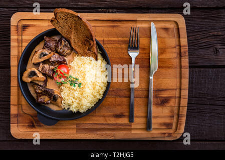 Vista superiore padella con saporite carni bovine stroganoff con funghi e il couscous è servita nel ristorante con coltello e forchetta Foto Stock