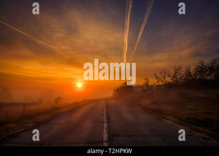 Sunrise visto da una strada di campagna con spaccature durante la stagione autunnale con drammatica nebbia e nuvole shot in Romania Foto Stock