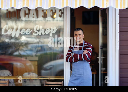 Un bel giovane coffee shop proprietario in piedi all'ingresso del suo negozio. Coffee shop iscrizione sulla finestra del negozio. Egli è un fiero proprietario del negozio Foto Stock