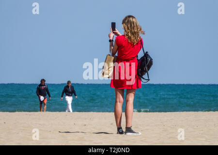 Valencia spiaggia Malvarrosa, una giovane donna in tuta rossa fare una foto su cellulare, vista posteriore Spagna vista mare Valencia spiaggia turisti Foto Stock