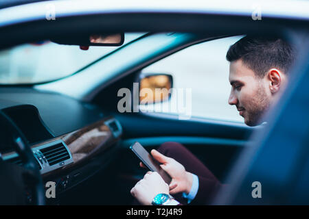 Giovane imprenditore guardando al suo cellulare stando comodamente seduti sul sedile anteriore nella sua limousine di lusso. Foto Stock