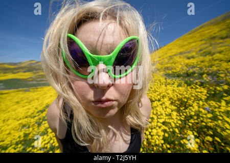 Spooky, creepy donna bionda che indossa il verde alien occhiali da sole in un campo di fiori selvatici goldfield in Carrizo Plain monumento nazionale durante il Californi Foto Stock