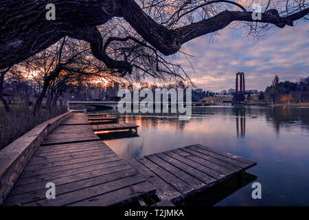 La mattina presto vista di un parco di Bucarest con un albero in primo piano e un monumento in background e le nuvole del cielo Foto Stock