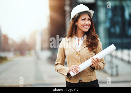 Ritratto di giovane donna ingegnere con casco bianco e blueprint nelle sue mani al sito in costruzione. Foto Stock