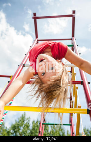 Elementare di età ragazza pendente da una giungla palestra (barre di scimmia o telaio di arrampicata) giocando in un parco giochi - sicurezza bambini o rischioso concetto di riproduzione Foto Stock