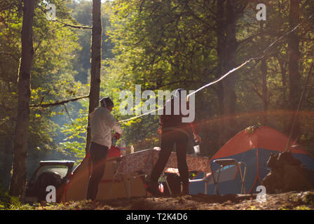 Una scena di un accampamento nel profondo la naturale vecchio foresta con alti alberi verdi e tende e persone e riflessi di luce solare attraverso Foto Stock