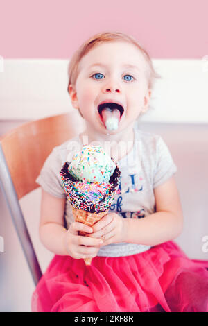 Carino divertente bionda caucasica babyl ragazza bambino con gli occhi blu di mangiare leccare il gelato in gran cono di cialda con spruzzino e che mostra la linguetta. Felice chi Foto Stock