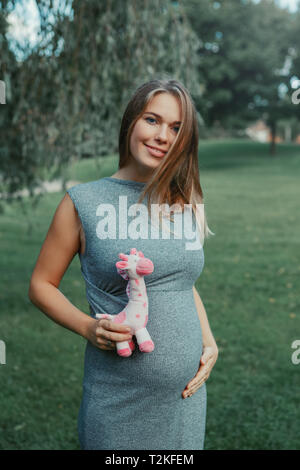 Ritratto di white Caucasian donna incinta di toccare la sua pancia e azienda rosa farcite giocattolo di peluche giraffa. Sorridendo ci aspettavamo femmina giovane madre in par Foto Stock