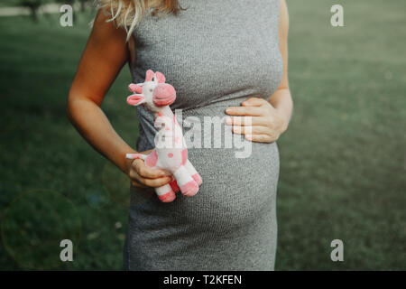 Closeup corpo del Caucaso in bianco donna incinta di toccare la sua pancia e azienda rosa farcite giocattolo di peluche giraffa. Ci aspettavamo femmina giovane madre in posa Foto Stock