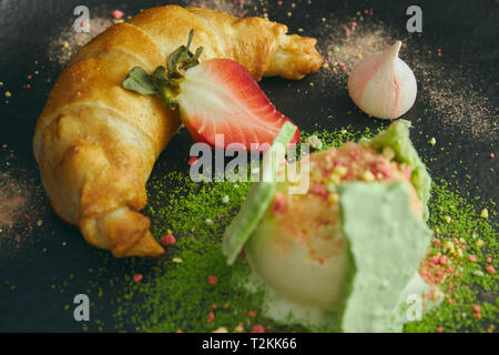 Fetta di strudel di mele con una pallina di gelato alla vaniglia su una piastra nera, su di un legno nero tabella, decorata con meringa e fragole. close-up Foto Stock