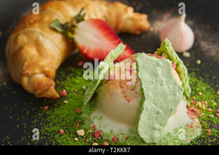 Fetta di strudel di mele con una pallina di gelato alla vaniglia su una piastra nera, su di un legno nero tabella, decorata con meringa e fragole. close-up Foto Stock