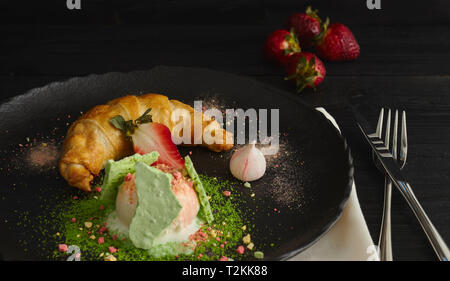 Fetta di strudel di mele con una pallina di gelato alla vaniglia su una piastra nera, su di un legno nero tabella, decorata con meringa e fragole. close-up Foto Stock