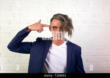 Ritratto di elegante imprenditore con ricci capelli lunghi in bicchieri guardando la telecamera su sfondo bianco. Mantiene il suo dito come una pistola a testa. Cervello bl Foto Stock