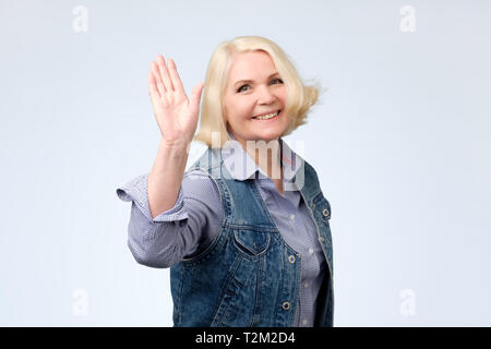 Bionda donna anziana in abiti casual agitando la mano dicendo hi, saluto i suoi amici. Foto Stock