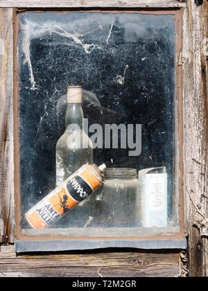 Uno sguardo attraverso la finestra di un antico giardino capannone abbandonato con bottiglie e lattine visto attraverso la ragnatela ricoperto di vetro. Foto Stock