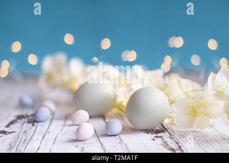 Colore naturale uova di Pasqua, malto caramella ricoperta di uova di cioccolato e fiori su un rustico di legno bianco tavola contro uno sfondo blu. Foto Stock