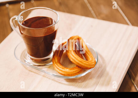 Un bicchiere di vetro con cioccolata calda e due frittelle spagnola servita su una tavola di legno Foto Stock