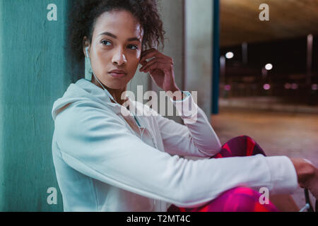 Sportive adegua la sua cuffia durante una pausa di allenamento. Guida femmina l'ascolto di musica in cuffia durante la pausa all'aperto nella notte. Foto Stock
