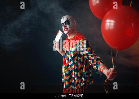 Brutto sanguinosa clown con carne cleaver detiene il pallone aerostatico, horror. L'uomo con il trucco in costume di carnevale, mad maniac Foto Stock