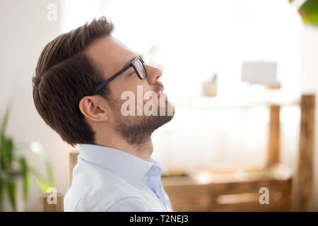 Close up attraente imprenditore millenario rilassarsi dopo il lavoro respirando aria fresca seduti in ufficio con gli occhi chiusi. Persona sognare thinkin Foto Stock