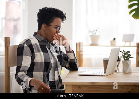 Esaurito imprenditrice africana in occhiali seduto alla scrivania in ufficio. Lavoratore o studente donna sbadigli coprono la bocca con la mano, si sente stanco di un Foto Stock