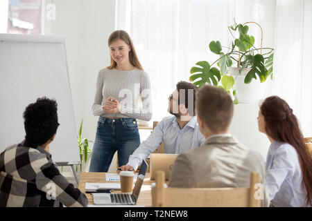 Diversi imprenditori durante il seminario seduto in ufficio. La millenaria leader femmina pullman in piedi vicino a whiteboard parlare di strategia di sviluppo a mu Foto Stock