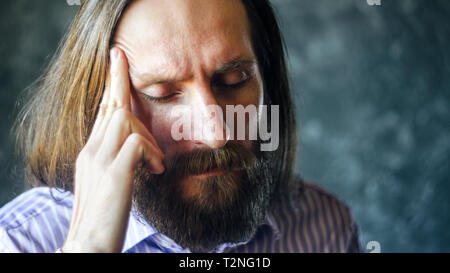 Ritratto di un uomo stanco con la barba e capelli lunghi con gli occhi chiusi strofinando il suo tempio di destra Foto Stock