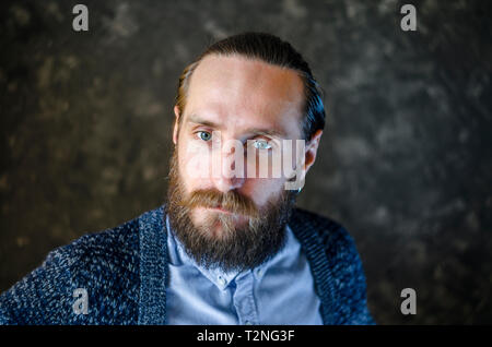 Ritratto di moda di un pensieroso uomo barbuto in una camicia blu e blu cardigan lavorato a maglia su sfondo grigio. Riflessività Concept Foto Stock