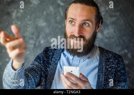 Uomo Barbuto hanno Business Meeting tramite chiamata video e Gesticulates durante la conversazione contro il muro grigio in un ufficio Foto Stock