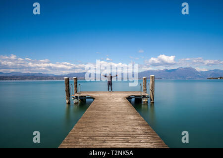 Uomo in piedi su un molo con le mani spalancate dal tranquillo lago di Garda, Italia Foto Stock