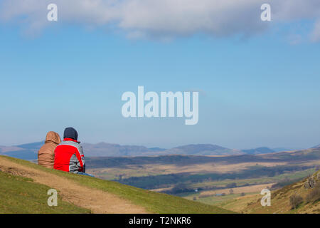 Vista posteriore del giovane fino una montagna nel Mid-Wales seduti sul pendio erboso, nella luce del sole, con lo sguardo al paesaggio spettacolare vista di fronte a loro. Foto Stock