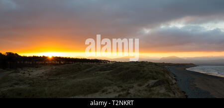 Vista panoramica di sunrise a Newborough Warren, Anglesey. Il sorgere del sole su Snowdonia montagne in distanza come dawn si rompe; nuvole scure, scuro litorale. Foto Stock