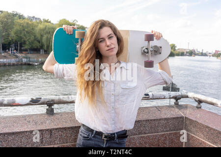 Giovane donna con lo skateboard in spalla, fiume in background, Berlino, Germania Foto Stock