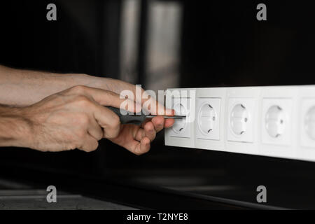 Close-up di un elettricista la mano con un cacciavite smontare un bianco presa elettrica su una nera parete di vetro. Foto Stock